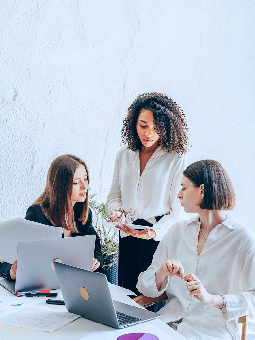 Three women are collaborating at a table with laptops and papers, discussing ideas in a bright, modern workspace.